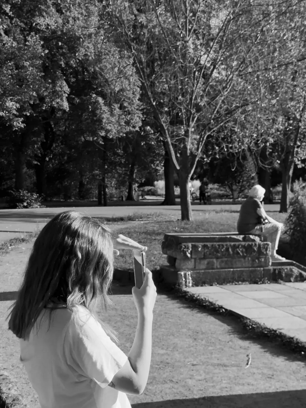 A young girl stands on a grassy area holding a phone, focused on taking a photo. In the background, an older woman sits on a stone bench under trees with bare branches, while sunlight filters through the foliage.
