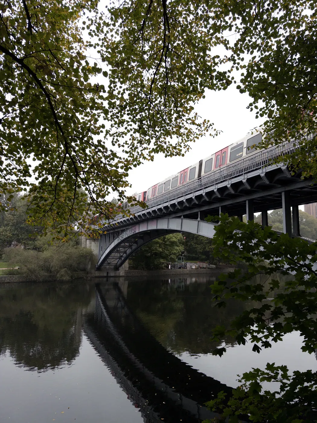 A train crosses a bridge above a calm river, surrounded by lush green foliage. The reflection of the bridge and train can be seen in the water below, creating a serene and picturesque scene.