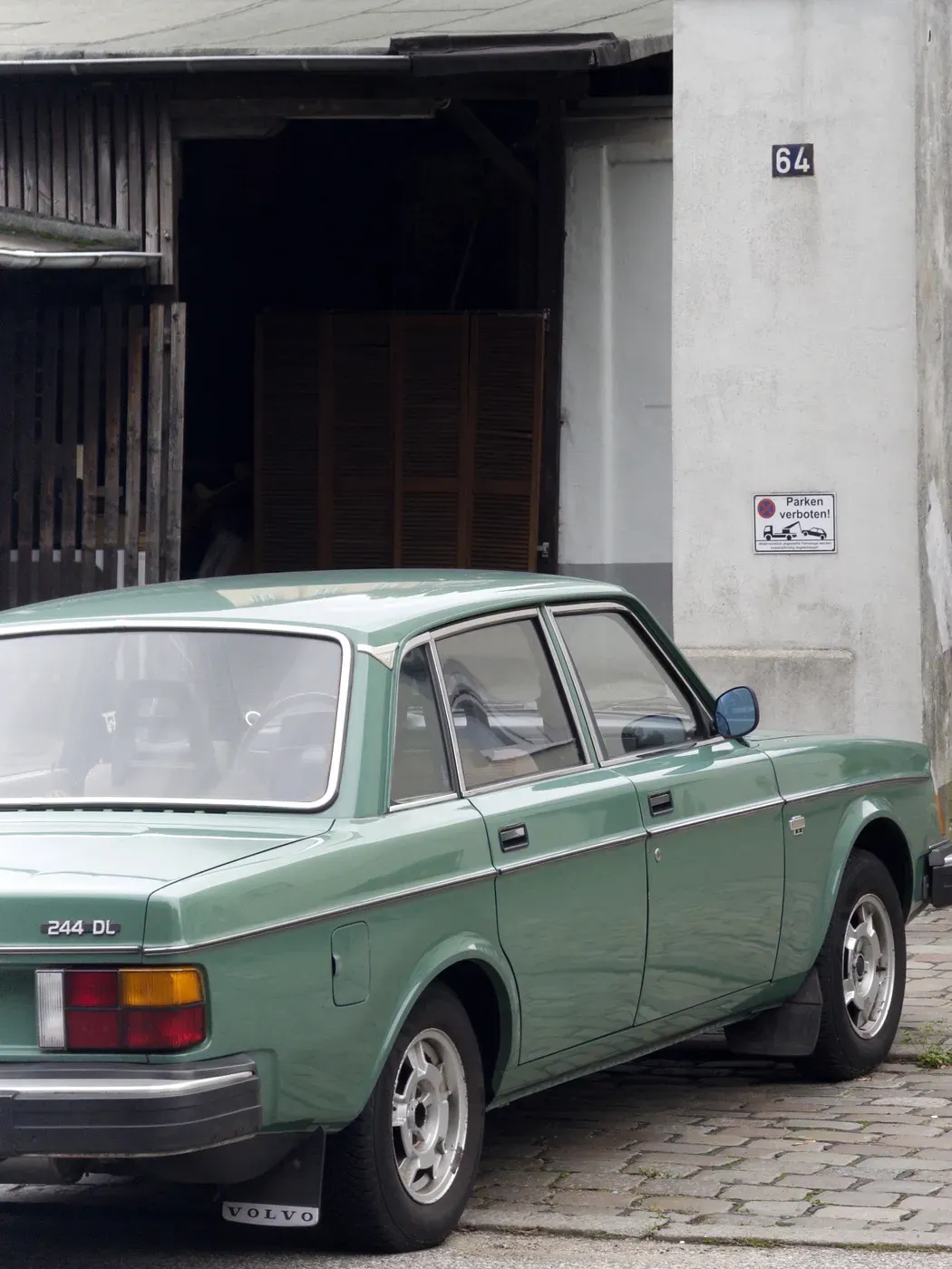 A vintage green Volvo 244 DL is parked next to a building with a wooden facade and a sign indicating a location. The scene captures a blend of rustic architecture and classic automotive design.