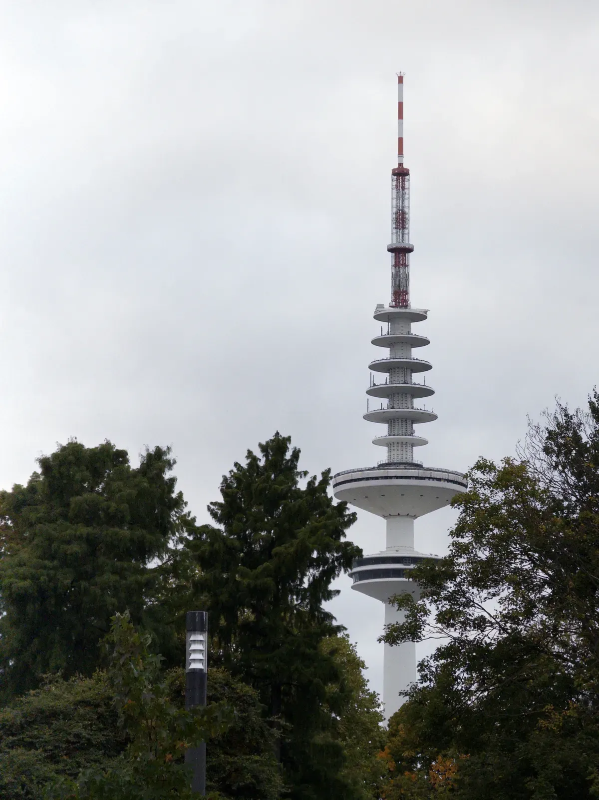 A tall communication tower rises amidst trees, featuring multiple circular platforms and a red and white antenna atop. The sky is overcast, creating a moody atmosphere.