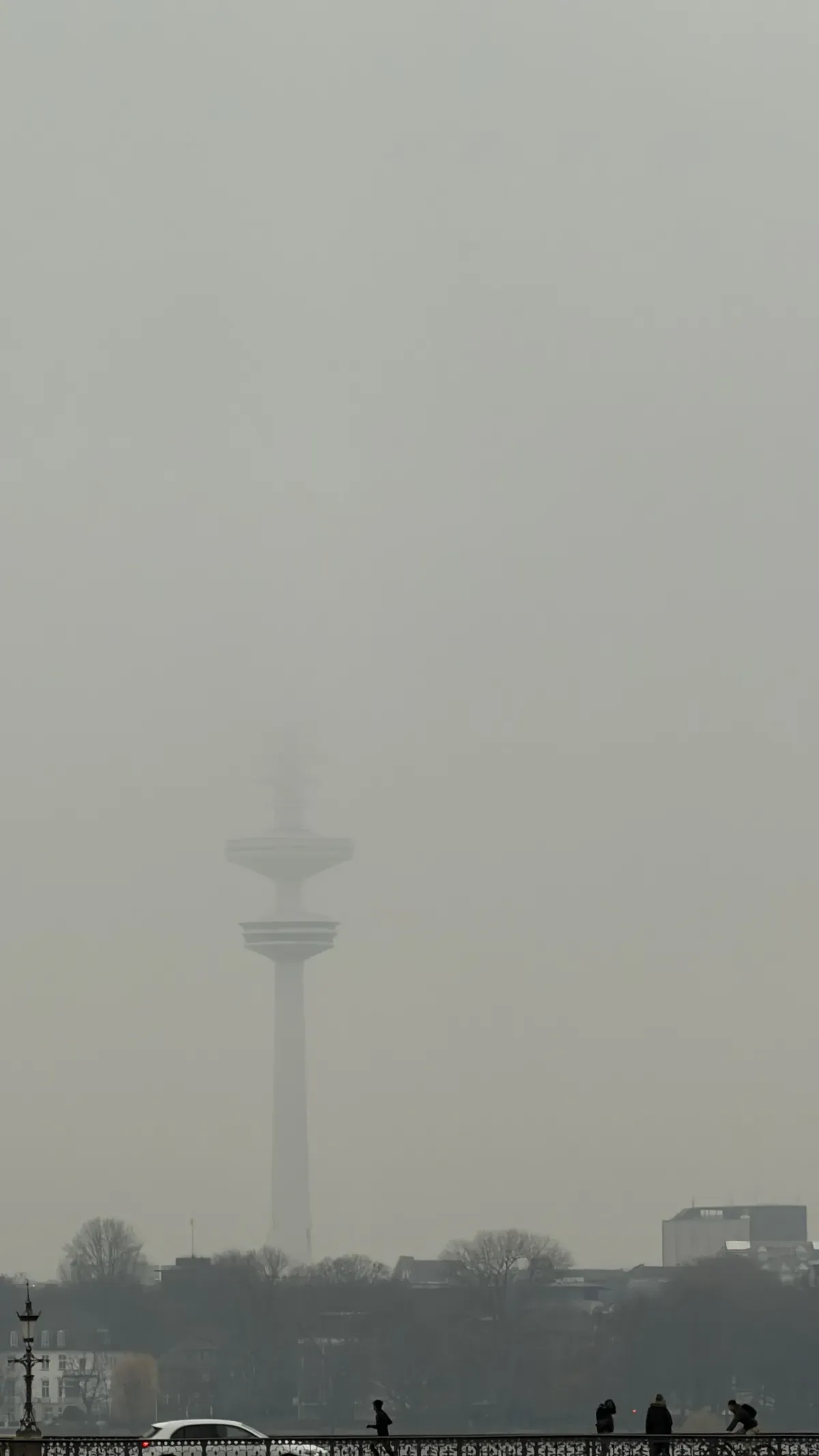 A tall tower is barely visible through thick fog, with only its top and antenna peeking out into the gray, misty atmosphere. The surrounding landscape is shrouded in haze, creating a subdued and mysterious ambiance.