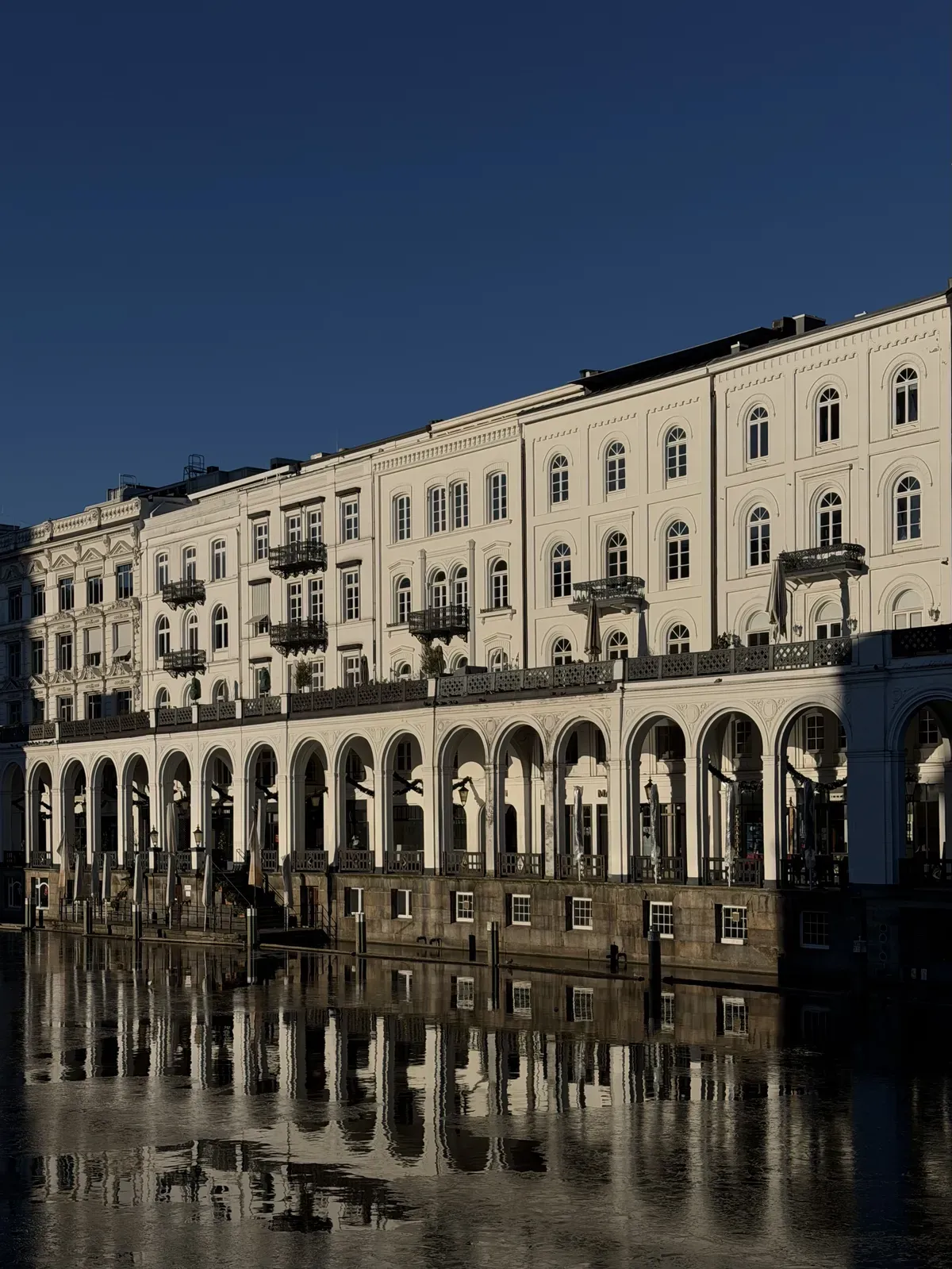 A white building with arched columns runs alongside a calm body of water, reflecting its structure. The scene is illuminated by sunlight against a clear blue sky, highlighting the architectural details and symmetry.