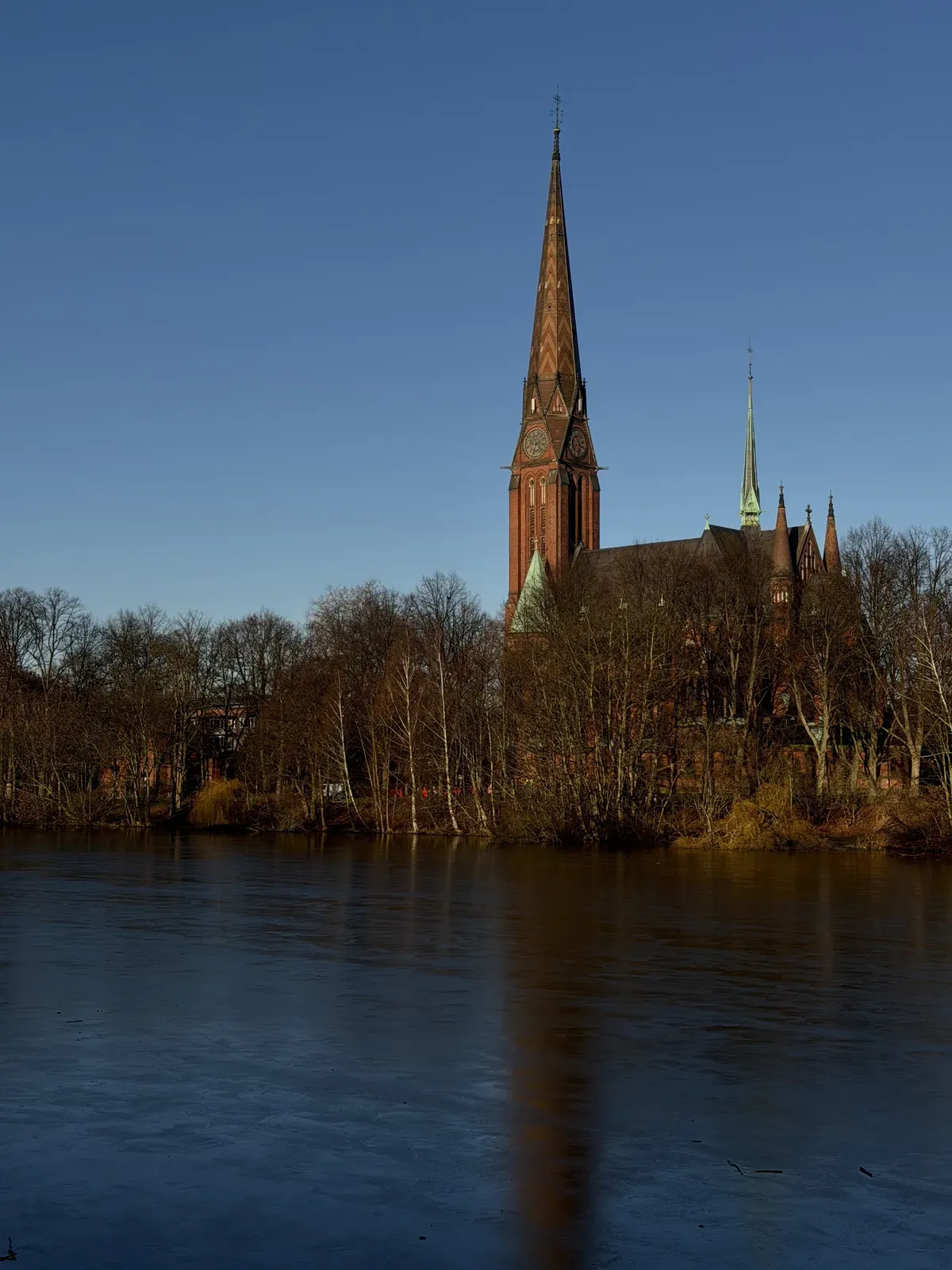 A tall, ornate church with a pointed steeple stands beside a calm body of water, reflecting its silhouette. Surrounding trees without leaves indicate a winter scene under a clear blue sky.