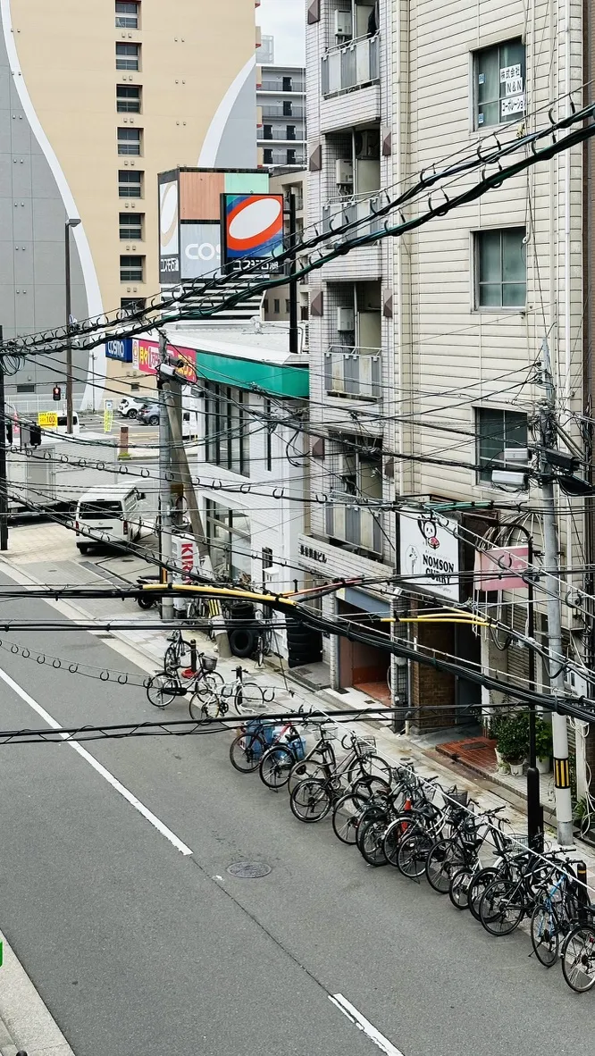A street scene featuring a row of parked bicycles along the side of the road, with buildings and power lines in the background. A gas station sign is visible in the distance, along with various storefronts.