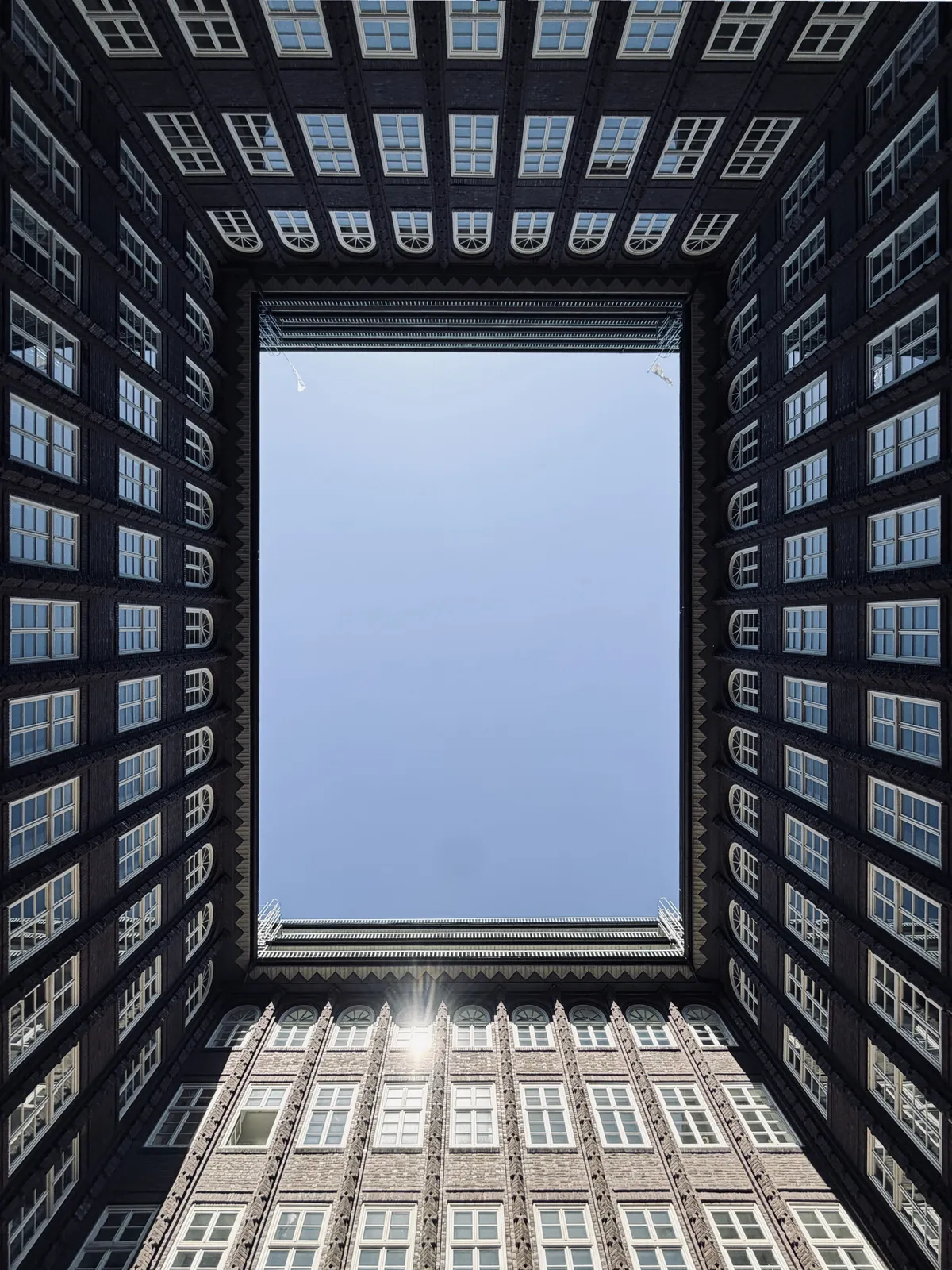 A view looking up through the inner courtyard of a building, framed by brick walls and rows of windows. The clear blue sky is visible in the center, creating a geometric contrast with the architectural details of the surrounding structure.