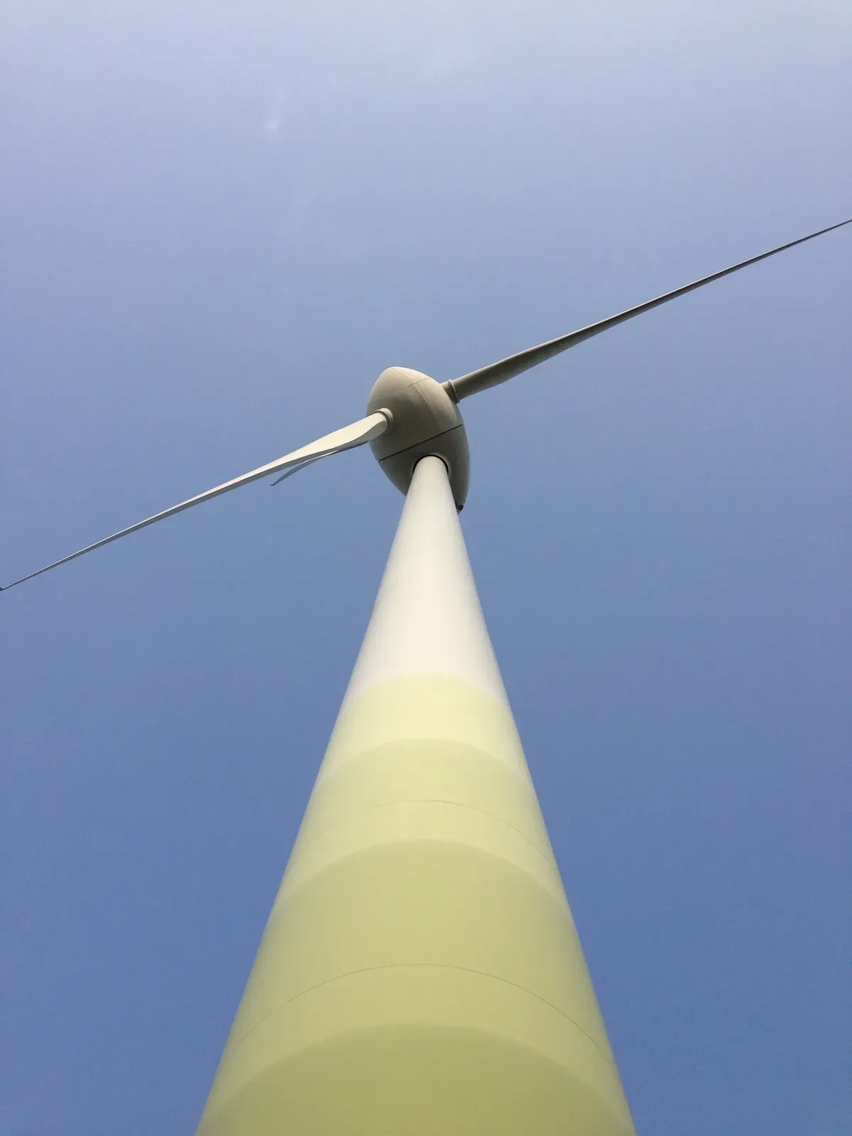 A view looking up at a tall wind turbine against a clear blue sky. The turbine's white tower is visible, with its three blades extending outwards at the top.