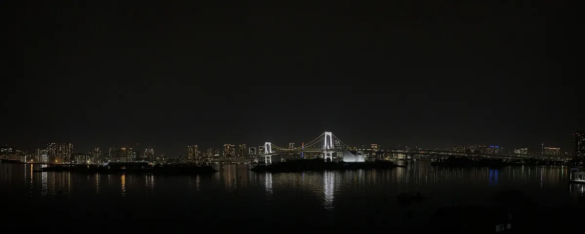 A panoramic night view of a city skyline featuring a bridge illuminated with bright white lights, reflecting on the calm water below. The scene includes skyscrapers and buildings with various colored lights in the background.