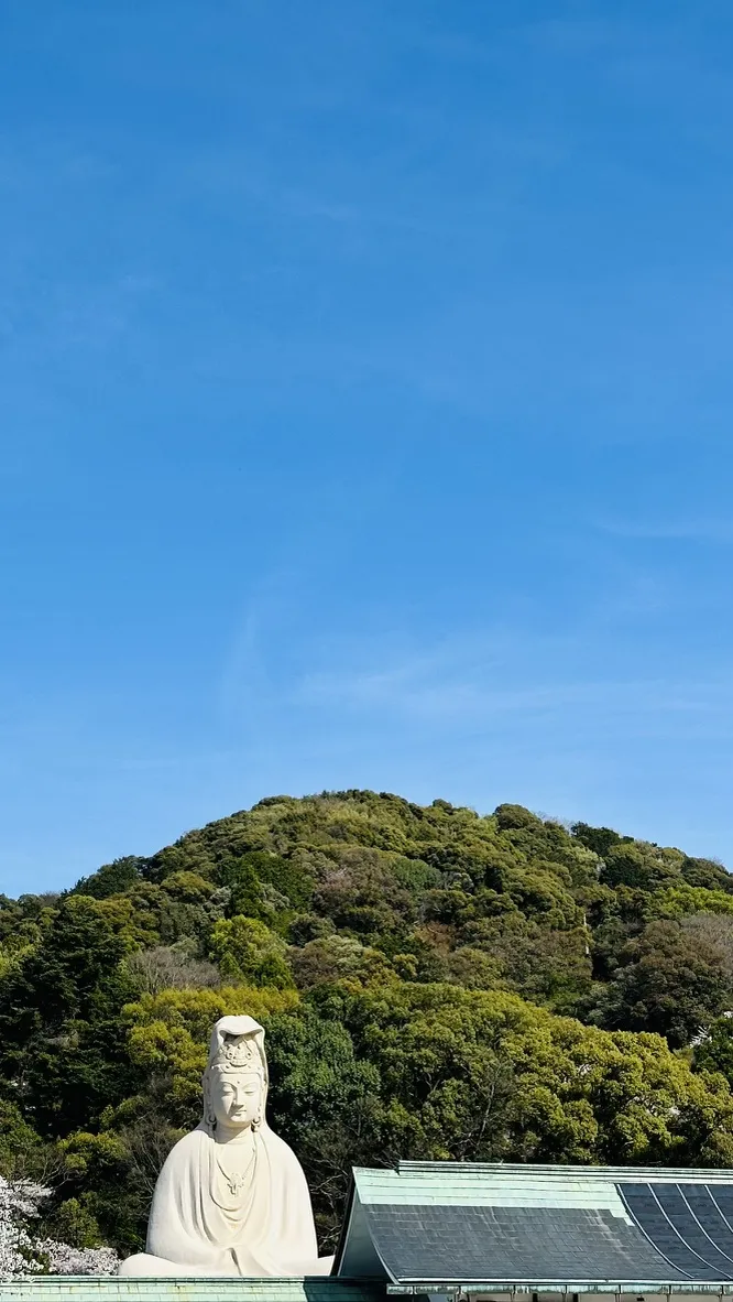 A large white statue of a seated figure is positioned in the foreground, with lush green hills rising behind it under a clear blue sky. The statue's serene expression contrasts with the vibrant natural landscape surrounding it.