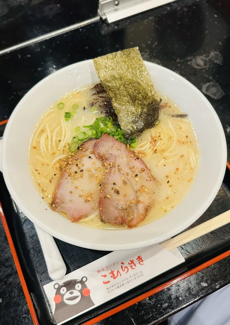 A bowl of ramen featuring a creamy broth topped with slices of pork, green onions, and a piece of seaweed. The bowl is set on a black surface with a small card featuring a bear character.