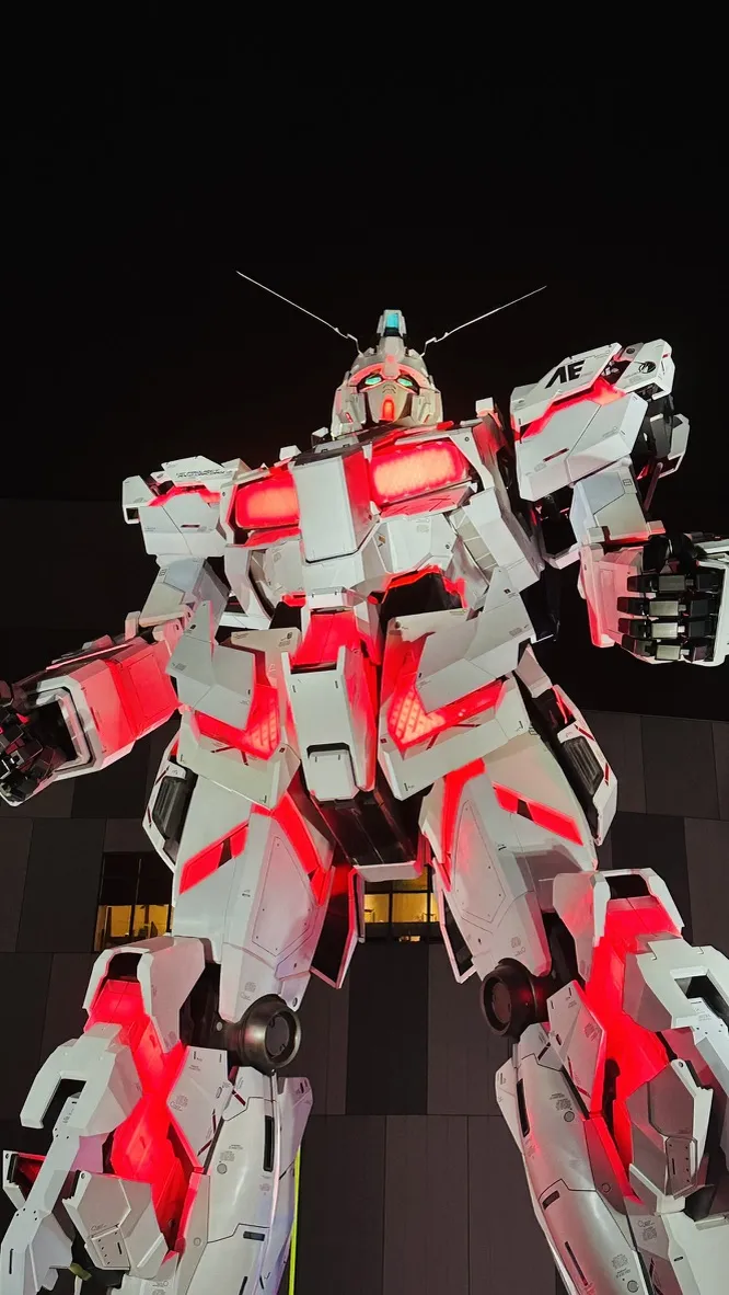 A large, humanoid robot sculpture stands tall against a dark night sky, illuminated by bright red lights on its chest and limbs. The robot has a sleek, white exterior and appears to be in a dynamic pose, with one arm raised.