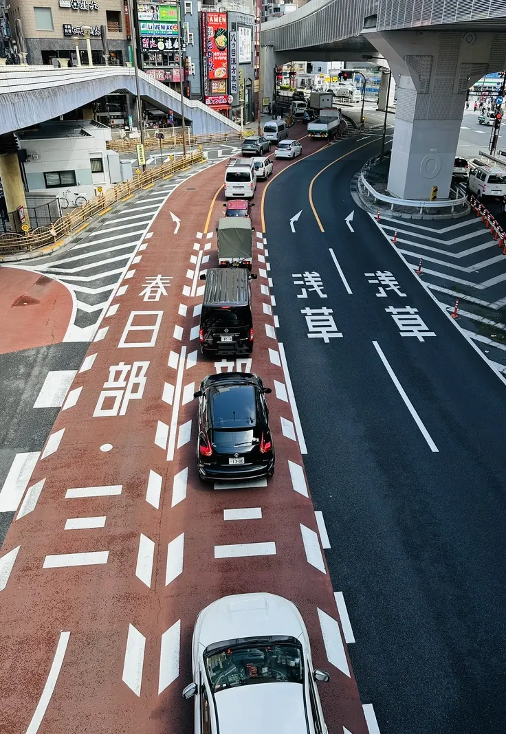 Traffic is seen on a two-lane road with clearly marked lanes merging into a curved path. Multiple vehicles, including cars and trucks, are waiting in line, while colorful advertisements and buildings are visible in the background.