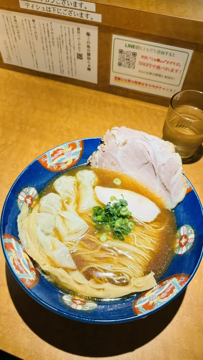 A bowl of ramen featuring yellow noodles, slices of pork, and a soft-boiled egg, garnished with chopped green onions. The broth appears rich and dark, with some wontons visible alongside the noodles.