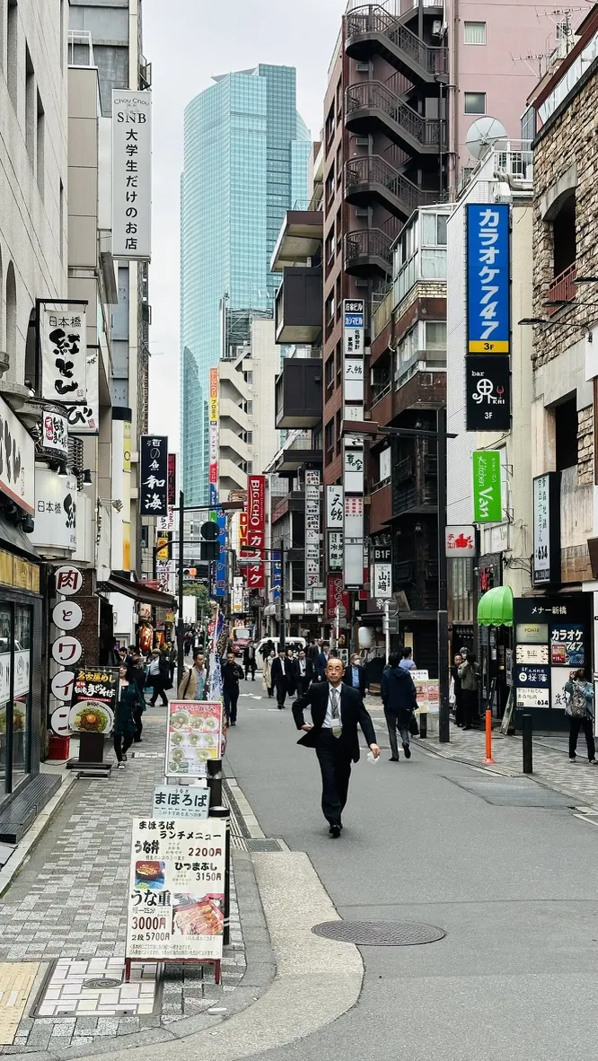 A bustling city street lined with various shops and restaurants, featuring bright signboards in multiple languages. In the foreground, a businessman walks briskly, while pedestrians can be seen along the sidewalk, amidst modern and traditional architecture.