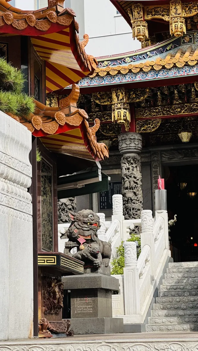 A decorative temple entrance features ornate rooftops with colorful patterns and intricate carvings. In the foreground, a stone statue of a mythical creature stands guard beside a staircase leading to the temple.