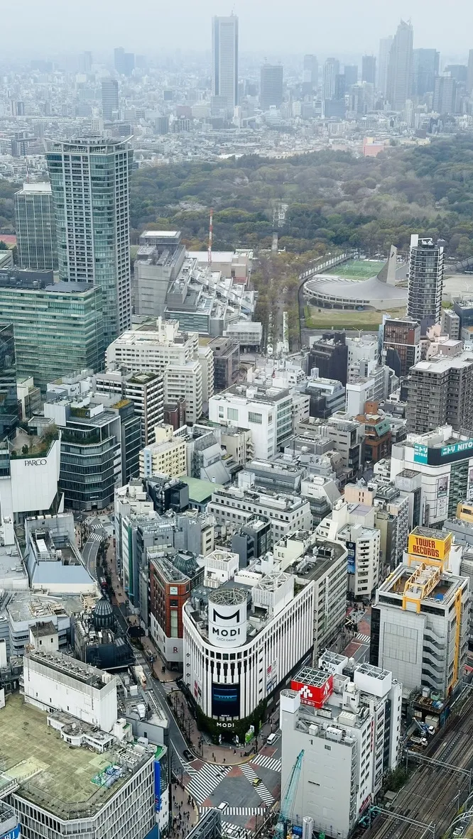 Aerial view of a bustling urban landscape featuring a mix of modern skyscrapers and a dense collection of buildings, with a prominent white structure labeled 'MODI' at the center. In the background, a sprawling green park contrasts with the city's concrete structures, and a distant skyline is visible under a cloudy sky.