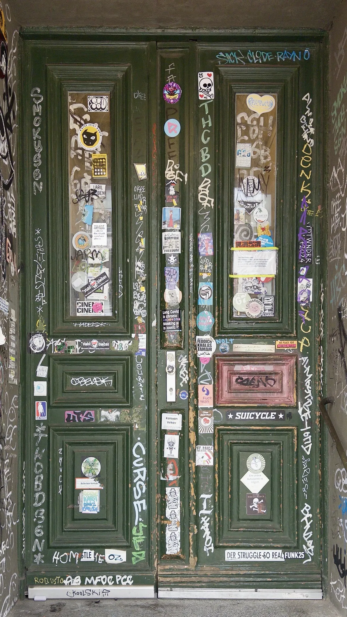 A green wooden double door covered in a variety of colorful stickers, graffiti, and tags. The door features two glass-paneled sections at the top and intricate wooden carvings.