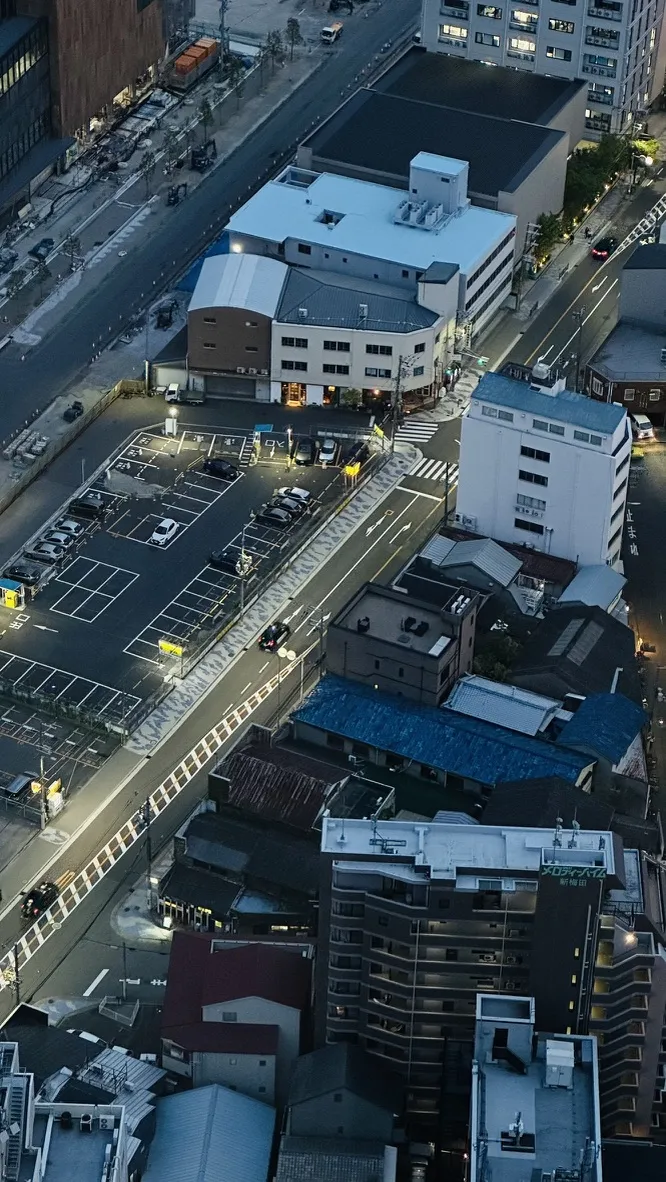 Aerial view of a city street at dusk, showcasing a mix of residential and commercial buildings. The scene includes parked cars in a lot and illuminated streetlights casting a warm glow on the roadway.