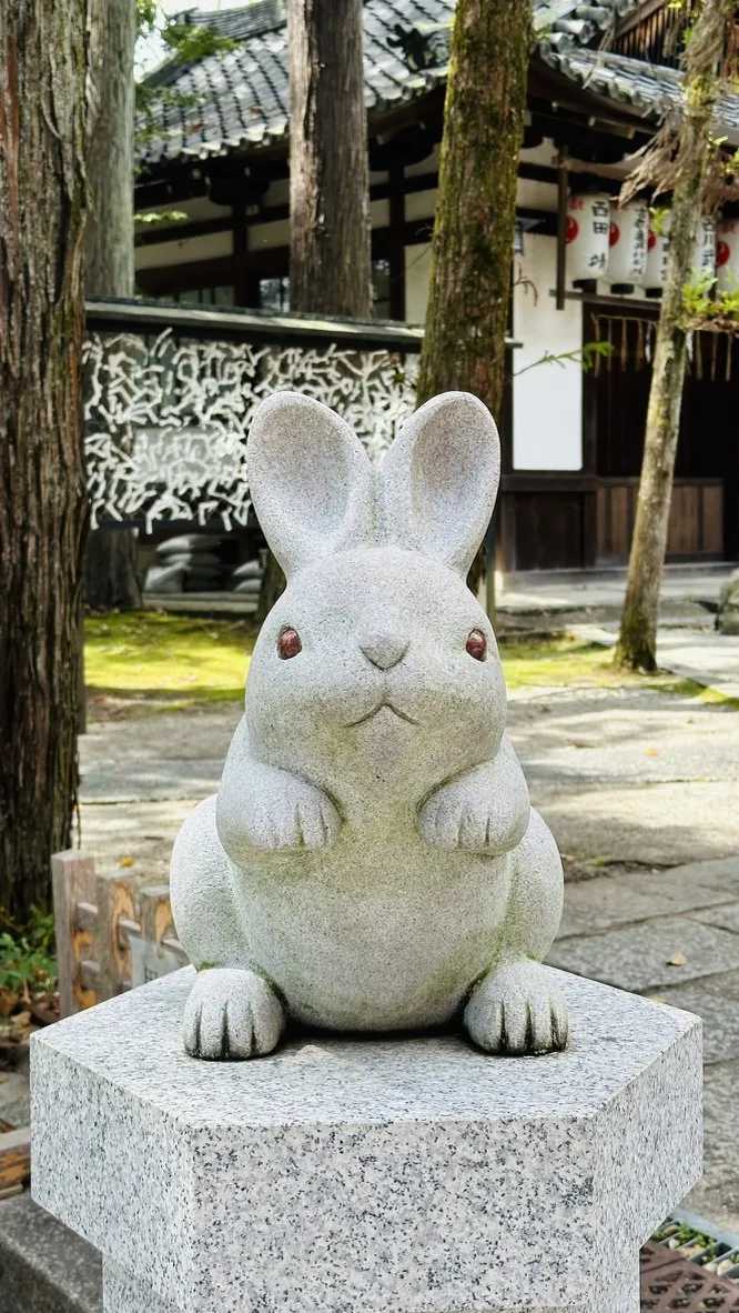 A stone sculpture of a sitting rabbit with large ears and small, round eyes is perched on a square pedestal. The background features a traditional Japanese architecture and trees, suggesting a serene outdoor setting.