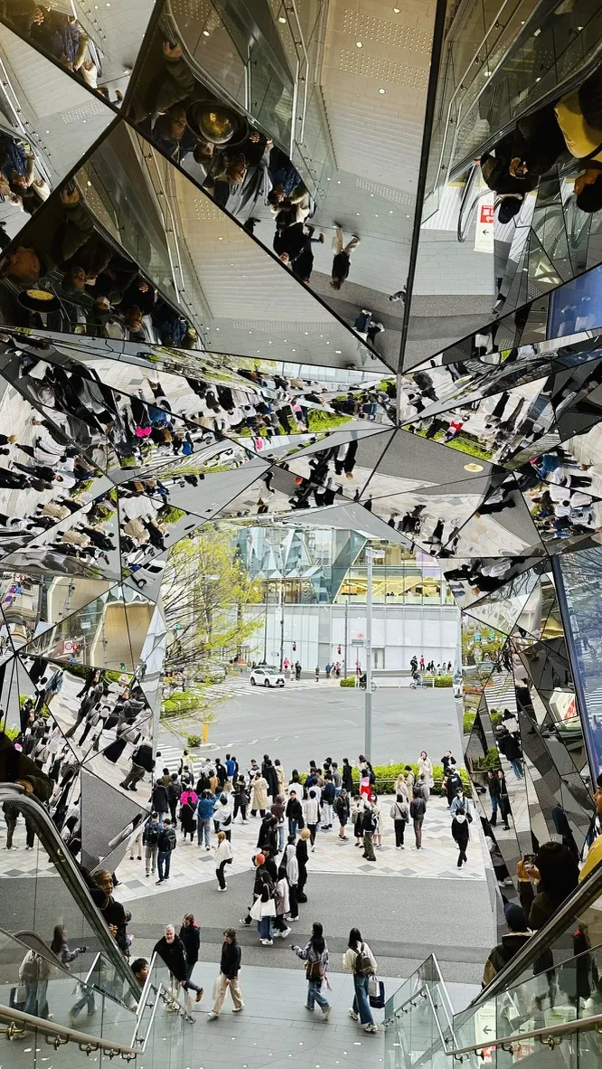 A spiral staircase with mirrored panels reflects the bustling activity of a busy urban area below. People are walking along the ground, while the design creates an intricate pattern of reflections and perspectives.