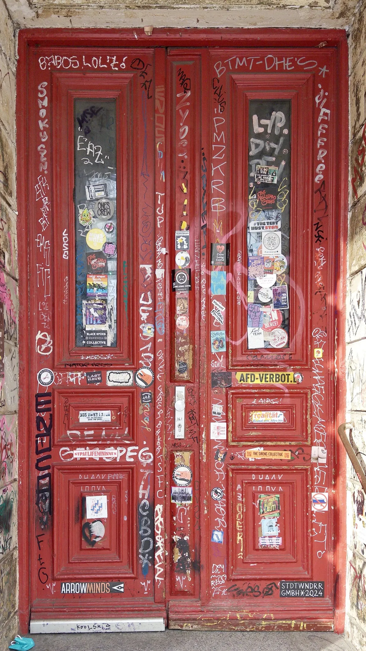 A pair of red double doors covered in a variety of colorful graffiti, stickers, and tags, showcasing an urban art scene. The doors have intricate designs and are set against a stone wall, adding to the vibrant display of street art.