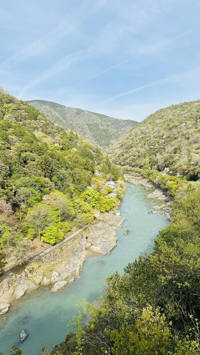 A serene river flows through a lush green canyon, bordered by steep, rocky hills. The water reflects a light blue sky, with soft clouds and a tranquil atmosphere.