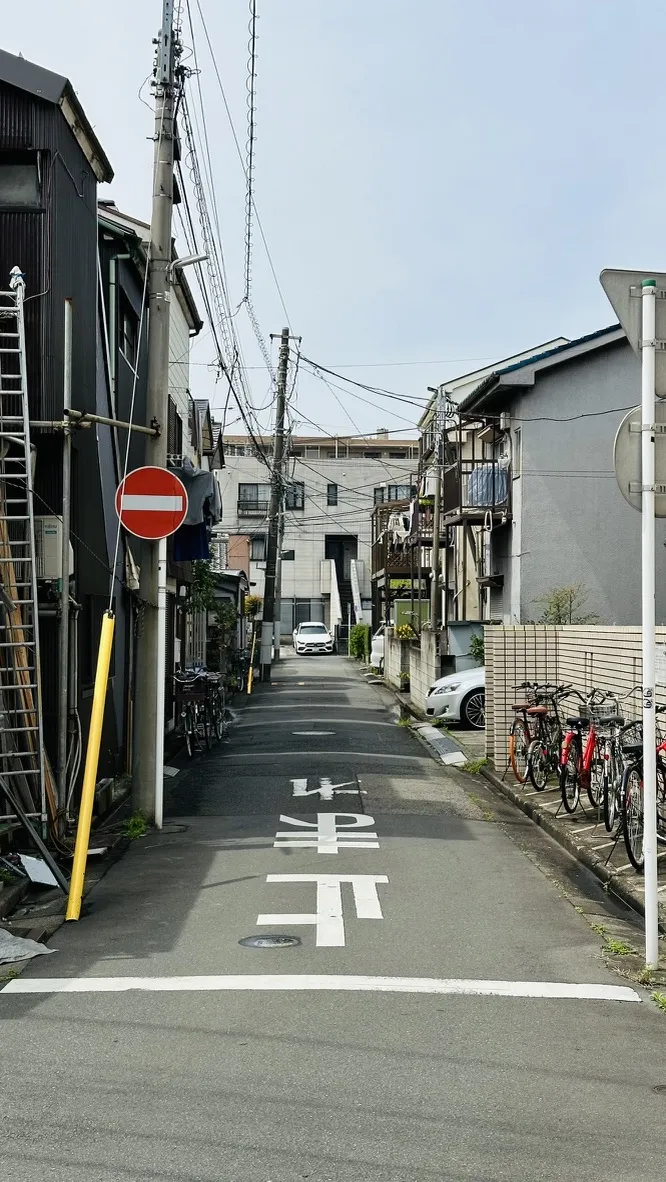 Narrow street lined with residential buildings and parked bicycles, featuring a no-entry road sign and power lines overhead. A white car is visible at the end of the alleyway.