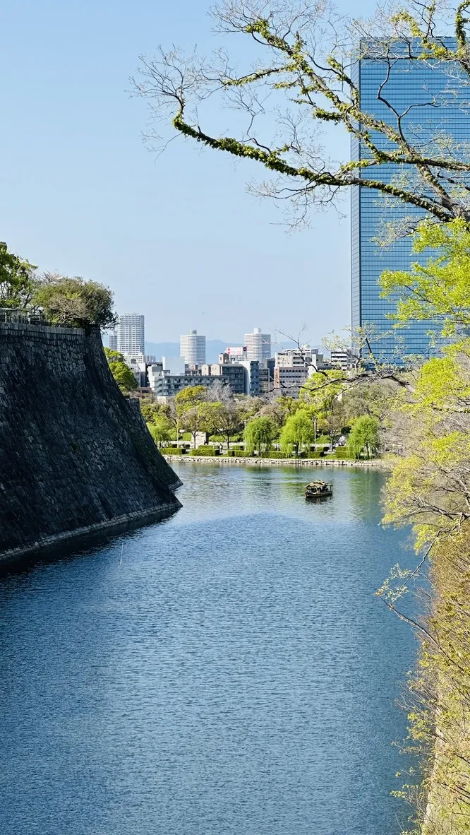 A serene river flows between the stone walls of a castle, with lush greenery lining the banks. In the background, modern skyscrapers rise against a clear blue sky.