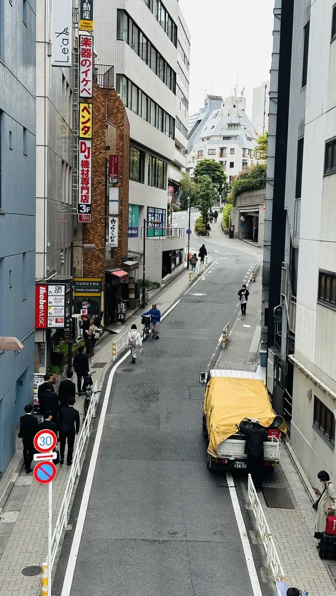 A narrow urban street in a city, featuring a mix of commercial buildings and a sloped road. Pedestrians walk along the sidewalk, while a yellow-covered truck is parked near the side, with traffic signs visible indicating a speed limit.