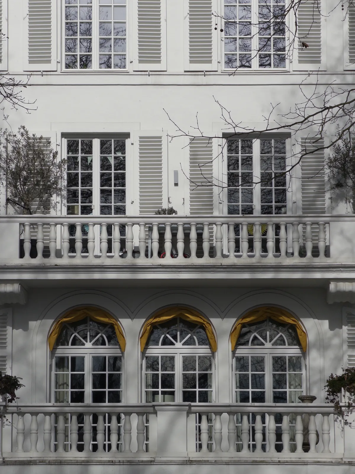 A white building features a symmetrical facade with multiple windows adorned with shutters. The upper level has decorative balcony railings, while the lower level showcases arched window frames with yellow drapes.