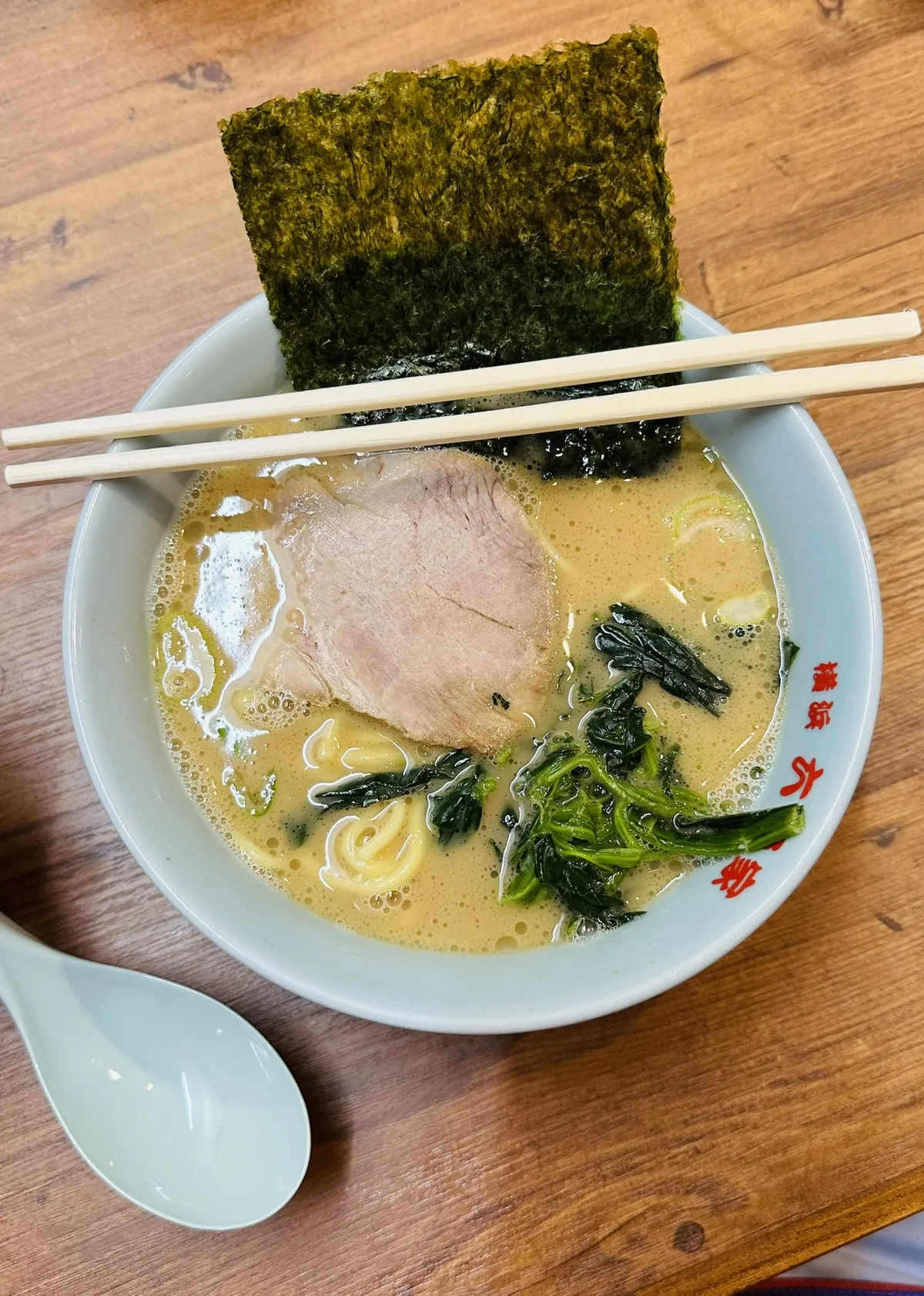 A bowl of ramen soup with a creamy broth features slices of pork, leafy greens, and seaweed on top. Two chopsticks rest on the edge of the bowl, and a spoon is placed beside it on a wooden table.