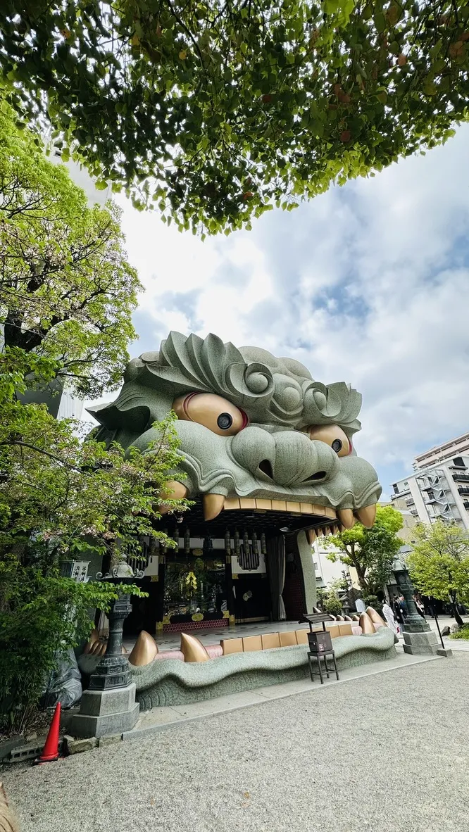 A large, decorative lion statue with exaggerated features dominates the scene, surrounded by trees and a stone pathway. The sky is partly cloudy, and the statue is part of a shrine structure, featuring traditional elements and a serene atmosphere.