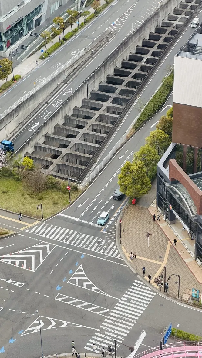 An aerial view of a city intersection featuring a roadway with distinct crosswalks, a concrete trench running parallel to the street, and surrounding green areas. Pedestrians can be seen walking along pathways, and modern buildings are in the background.