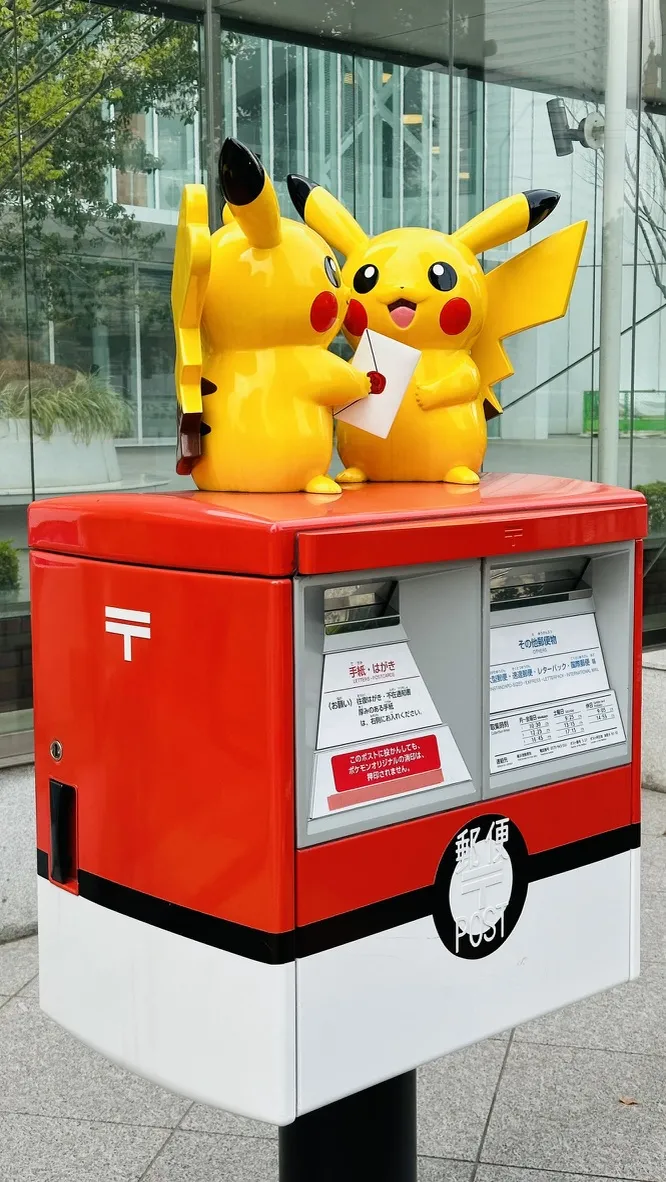 Two large Pikachu figurines are playfully positioned on top of a bright red Japanese post box, each holding a letter. The post box features a white emblem and is located in an outdoor setting with greenery in the background.