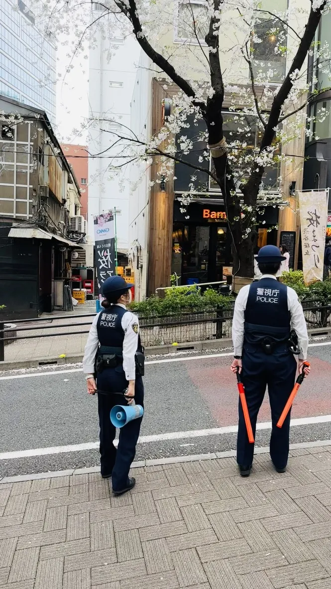 Two police officers in uniform stand on a city street, one holding a megaphone and the other holding a baton. Cherry blossom trees are visible in the background, along with storefronts and signage.