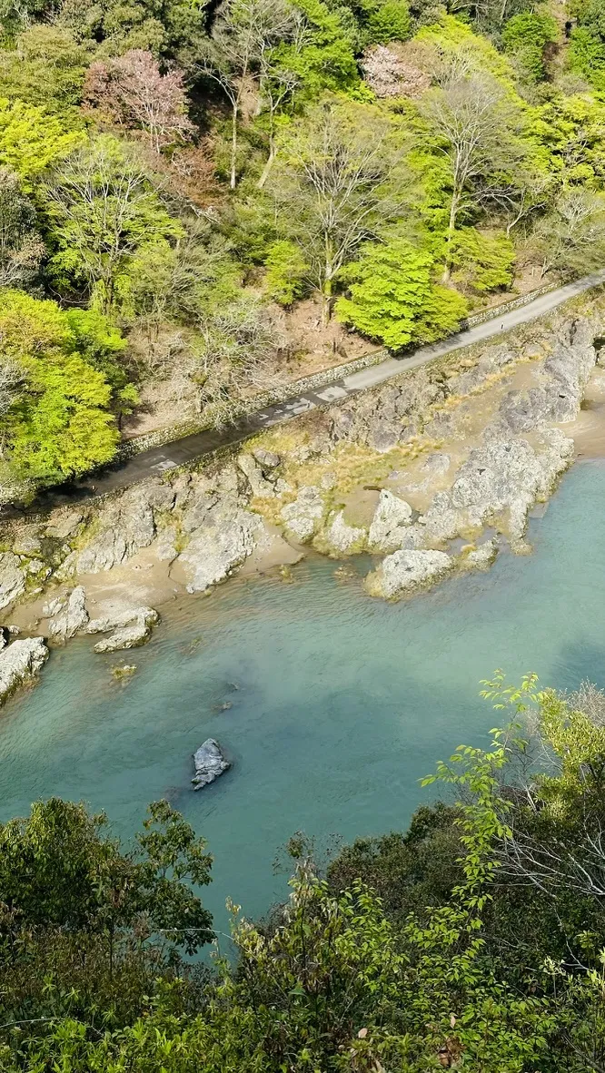 Aerial view of a winding river surrounded by lush greenery and rocky shores. The water is clear and reflects the natural landscape, while a road runs parallel to the riverbank.