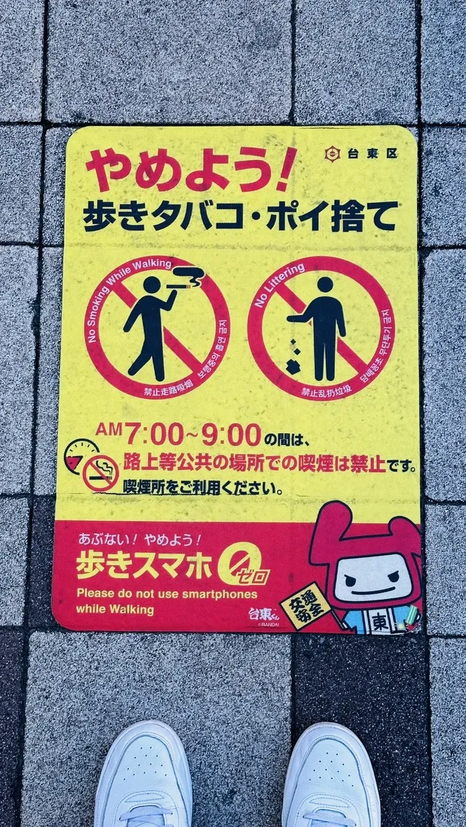 A colorful pavement sign in Japanese, featuring illustrations of a person walking and a person using a smartphone. It displays warnings against walking while smoking or using smartphones, indicating that these activities are prohibited in public places during specified hours.