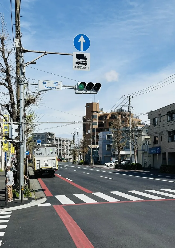 A street scene featuring traffic signals at an intersection. A truck is positioned near the crosswalk on a road marked with red lines, while buildings and trees line the sides of the street under a clear blue sky.
