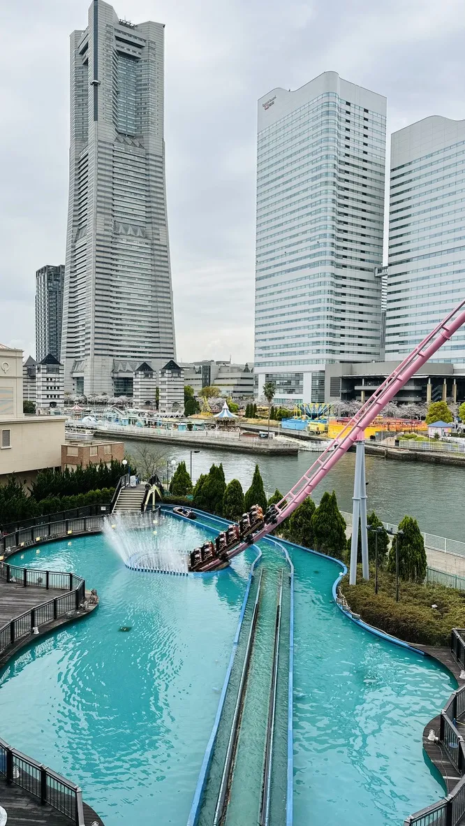 A water ride with a large splash features in the foreground, surrounded by lush greenery, with a modern city skyline in the background. High-rise buildings reflect a cloudy sky, and a calm waterway runs alongside the amusement park.