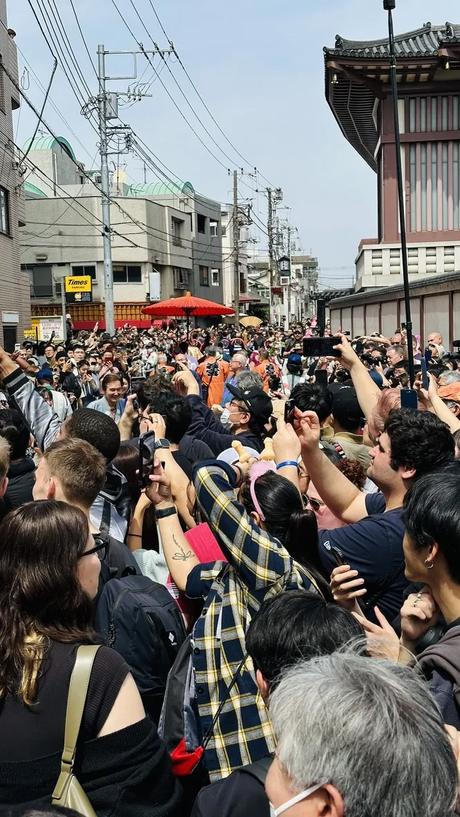 A large crowd of people is gathered in a bustling street, many holding up their phones to capture the moment. Buildings and power lines are visible in the background, creating a lively urban atmosphere.