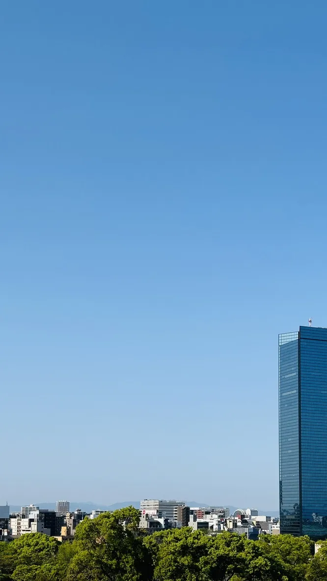 A clear blue sky stretches over a cityscape featuring a modern glass skyscraper on the right, surrounded by low-rise buildings and green trees in the foreground.