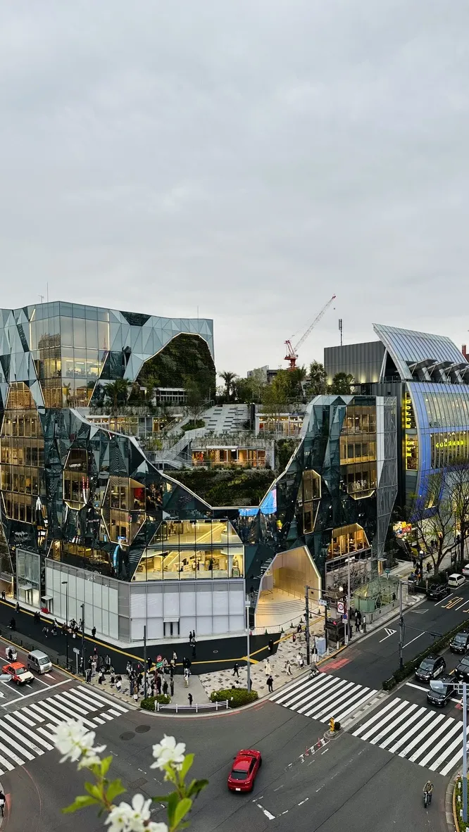 A striking modern building features a fragmented, reflective facade with angular designs. The scene includes a busy intersection below, with pedestrians crossing the street and vehicles passing by, set against a cloudy sky.