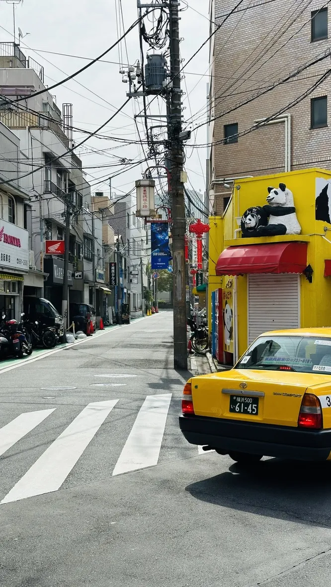 A yellow taxi is parked on a quiet street lined with small shops and buildings. Electrical wires crisscross overhead, and a store with panda decorations stands out on one side.
