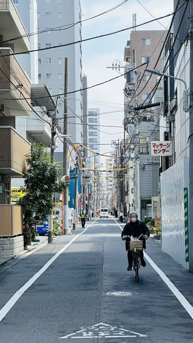 A narrow urban street lined with several buildings, featuring overhead power lines and a cyclist wearing a mask riding along the bike lane. In the background, taller buildings can be seen, and a sign in Japanese is visible on the right.