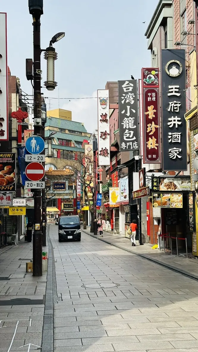A quiet street in a vibrant Chinatown area, lined with colorful signs in various languages advertising restaurants and shops. The road is mostly empty, featuring a few parked vehicles and traditional architecture in the background.