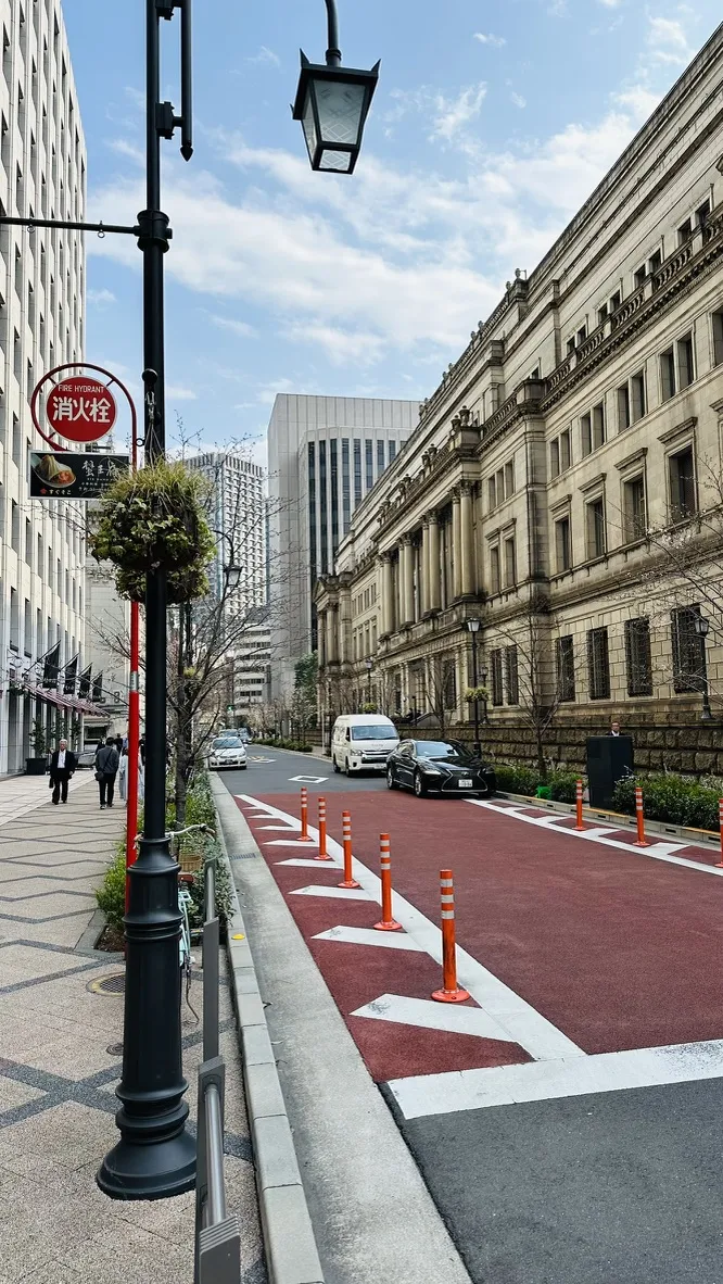 A street scene featuring a historic building on the right with classic architecture, alongside modern skyscrapers in the background. Traffic barriers in red and white line the road, which is partially marked with a fresh surface. A lamppost with a plant and a sign can be seen on the left.