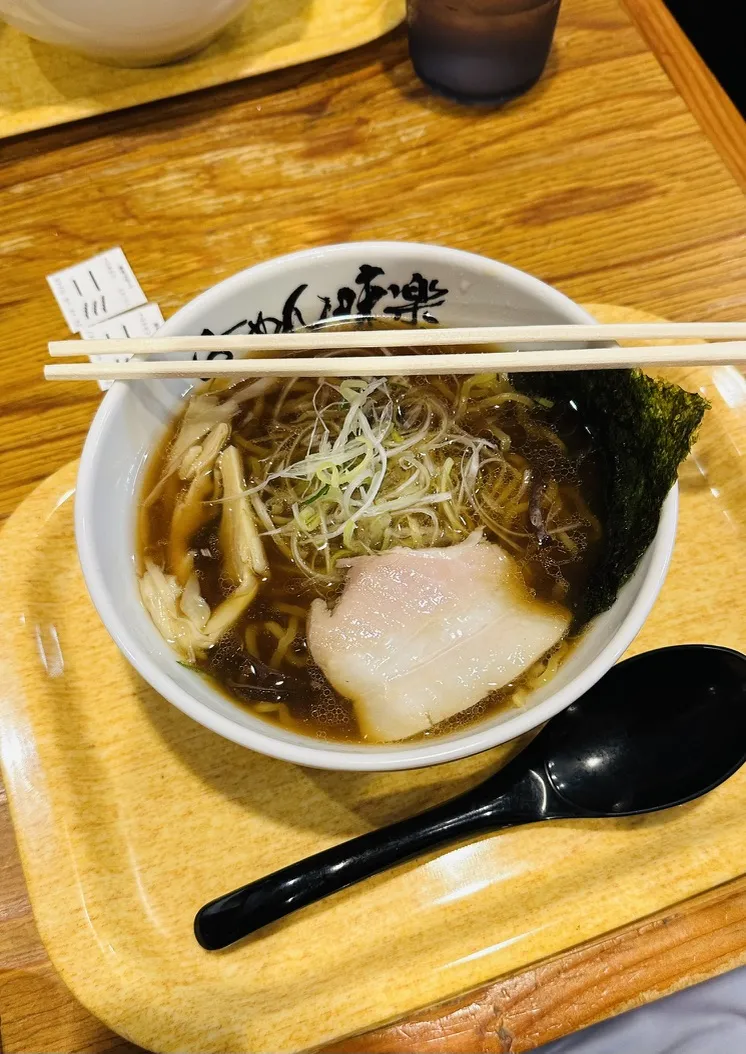 A bowl of ramen sits on a wooden table, featuring a rich broth topped with slices of pork, green onions, and a piece of nori seaweed. Chopsticks rest on the rim of the bowl, with a black spoon placed beside it.