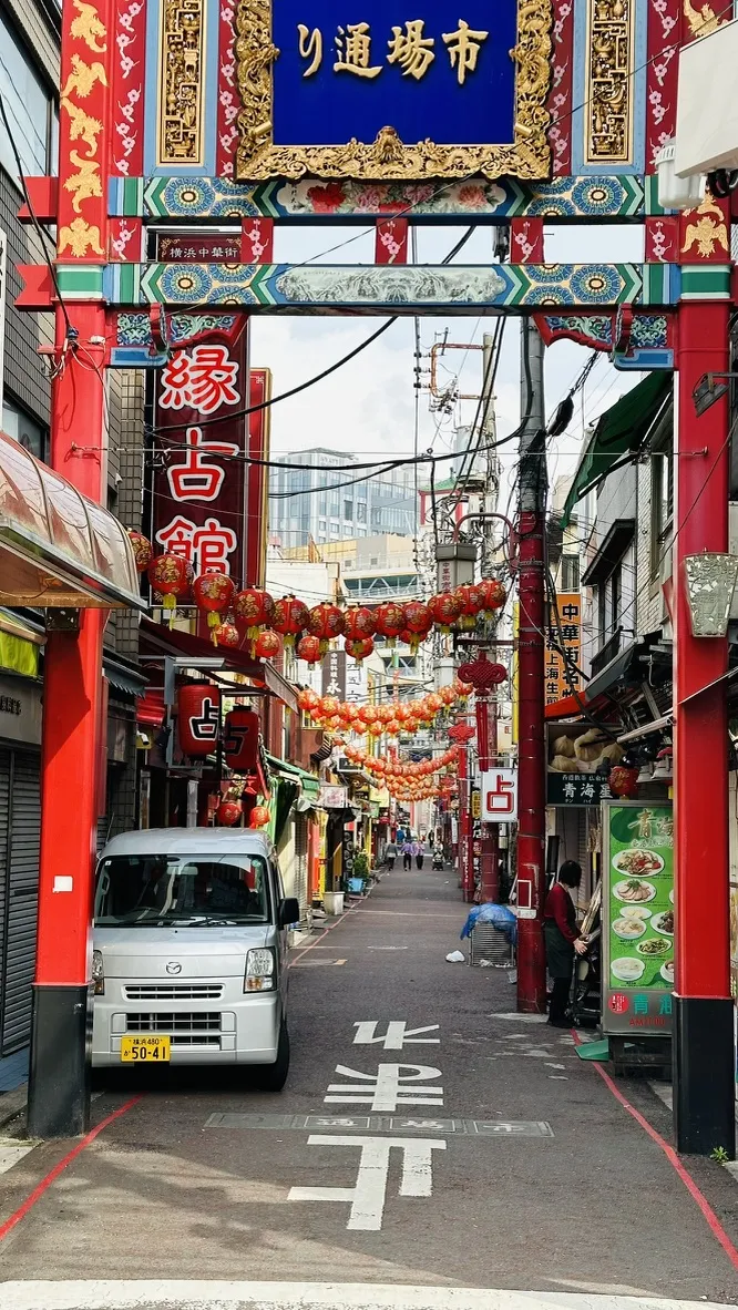 A narrow street lined with colorful red and green decorations, featuring hanging lanterns and signs in Japanese. A small white vehicle is parked on the left, while shops and eateries line both sides of the street.