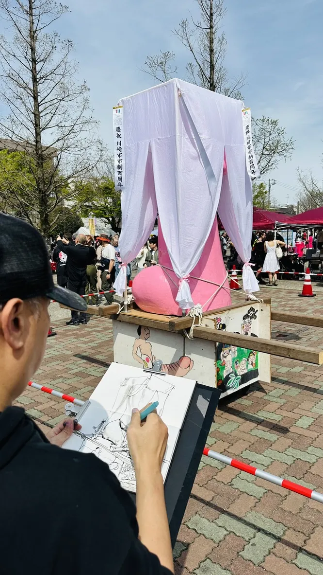A person sketches at an outdoor event, focusing on a large pink statue draped in white fabric. Surrounding the statue are festival tents, trees, and participants in traditional attire.