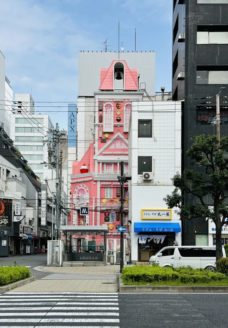 A colorful, pink-themed building features a whimsical design with a gabled roof and cartoon-like elements, set against a clear blue sky. Surrounding it are modern buildings and a crosswalk with greenery in the foreground.