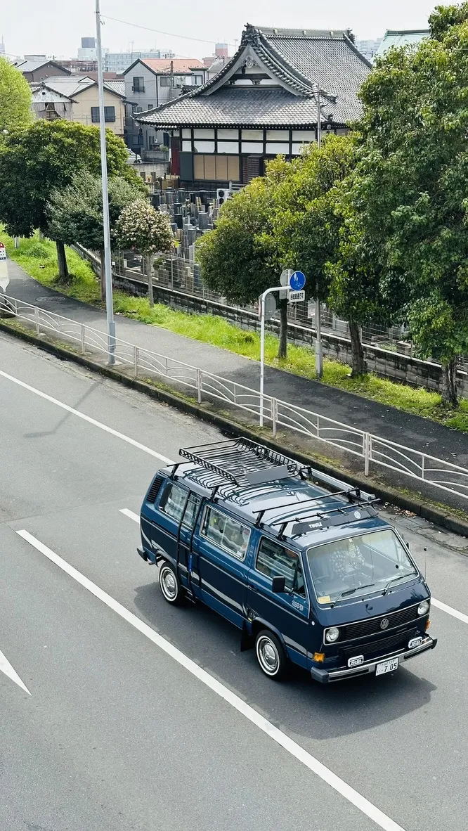 A vintage blue van drives along a quiet road, with a traditional Japanese building featuring a tiled roof and surrounding greenery visible in the background. Road signs and streetlights are also present along the roadside.